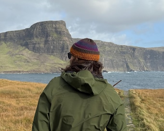 May include: A multi-colored knit beanie featuring horizontal stripes in shades of purple, blue, brown, and orange, worn with an olive green hooded jacket that has a small white reflective horizontal detail on the lower back.