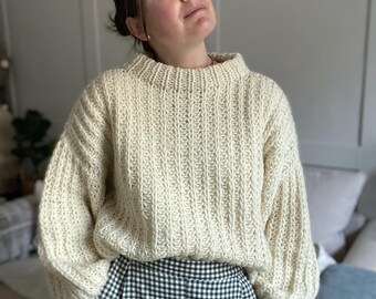 Könnte beinhalten: Ein cremefarbener Strickpullover mit Rippenstruktur und einem hohen Ausschnitt. Der Pullover wird über einer schwarzen und weißen Gingham-Hose getragen.