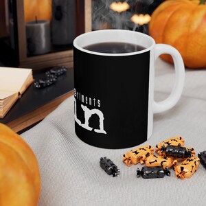 May include: A black coffee mug with a white rim and handle. The mug has the text "Experiments in" and a stylized "U.N." printed on it. The mug is sitting on a gray fabric surface with orange and black candy.