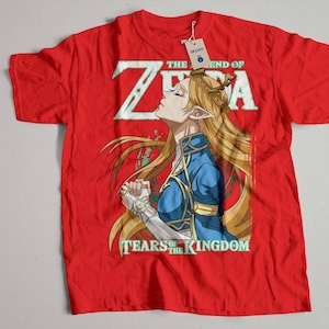 Puede incluir: Camiseta roja con un gráfico de Zelda de The Legend of Zelda: Tears of the Kingdom. El gráfico muestra a Zelda con el pelo largo y rubio y un atuendo azul y dorado. El texto "The Legend of Zelda: Tears of the Kingdom" está impreso en la camiseta.