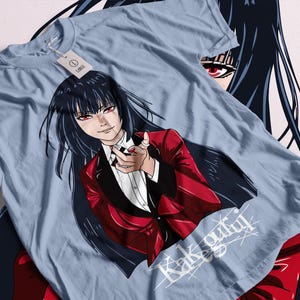 May include: A light blue t-shirt with a graphic of a woman with long black hair wearing a red blazer and a white shirt. The woman is smiling and holding out her hand. The text "Kakegurui" is written in white on the shirt.