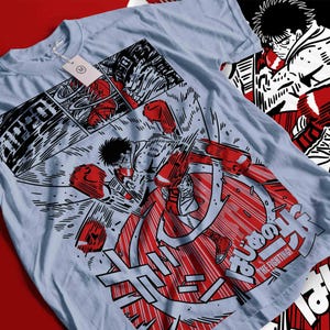 May include: Light blue t-shirt featuring a manga-style illustration of a boxer in action. The design incorporates red and white elements, with Japanese text and the words "THE FIGHTING!" The shirt has a tag attached.