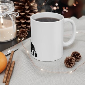 May include: White ceramic mug with a black silhouette of the Colosseum in Rome, Italy. The mug is filled with coffee and steam is rising from the top. The mug is sitting on a white surface with pine cones and string lights.