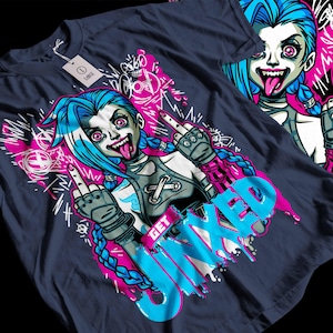 May include: Navy blue t-shirt featuring a cartoon illustration of a character with blue hair and pink eyes, sticking out their tongue and giving the middle finger. The words "GET JINXED" are in a dripping blue font. The design includes pink and white graffiti-style accents.