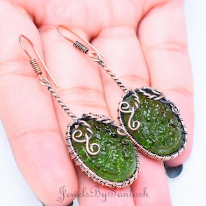 以下が含まれることがあります： 銅線で巻かれた緑色のガラス製のイヤリングのペア。イヤリングは楕円形で、繊細で複雑なデザインです。