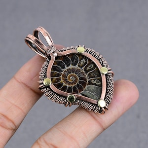 May include: A copper wire wrapped pendant featuring a fossilized ammonite with a brown and gold colour scheme. The pendant has a teardrop shape and is accented with small gold beads.