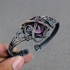 Pode incluir: Uma pulseira envolvida em fio de prata e cobre com uma gema roxa no centro. A pulseira é intrincadamente desenhada com espirais e curvas.