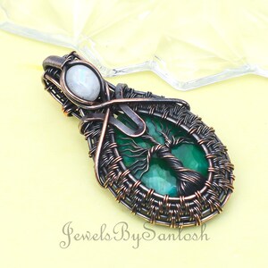 Op de afbeelding: Een hanger van koperdraad met een groene levensboomsteen met een witte maansteenaccent. De hanger is ovaalvormig en heeft een geweven draadontwerp.