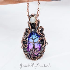 May include: A copper wire wrapped pendant featuring a purple and blue iridescent glass tree of life design. The pendant is accented with a white moonstone and hangs from a copper chain.