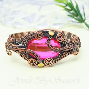May include: A handcrafted copper wire bracelet with a large, oval, pink gemstone at its center. The bracelet features intricate wirework with spiral accents and small brass-colored embellishments. The text "Jewels By Santosh" is at the bottom.