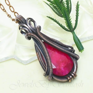 May include: A handcrafted pendant necklace featuring a large, faceted, ruby-red gemstone. The stone is encased in intricate wirework with copper and silver tones. The necklace chain is a delicate copper color. The words "Jewels By Santosh" are visible in the image.