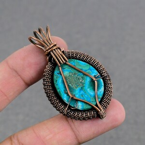 May include: A copper wire wrapped pendant featuring a turquoise blue and green gemstone with a swirling pattern. The pendant is held by a finger.