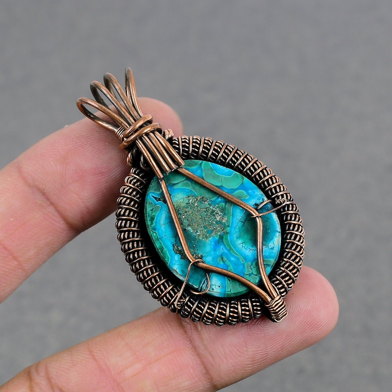 May include: A copper wire wrapped pendant featuring a turquoise blue and green gemstone with a swirling pattern. The pendant is held by a finger.