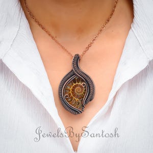 May include: A copper wire wrapped pendant necklace featuring a fossilized ammonite. The pendant is in the shape of a teardrop and has a woven wire design. The necklace is on a copper chain.