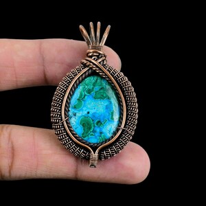 May include: A copper wire wrapped pendant featuring a green and blue gemstone. The pendant is oval shaped and has a detailed wire design.