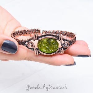 Puede incluir: Un brazalete envuelto en alambre de cobre con un cabujón de vidrio verde. El brazalete está tejido con un patrón de malla de alambre.