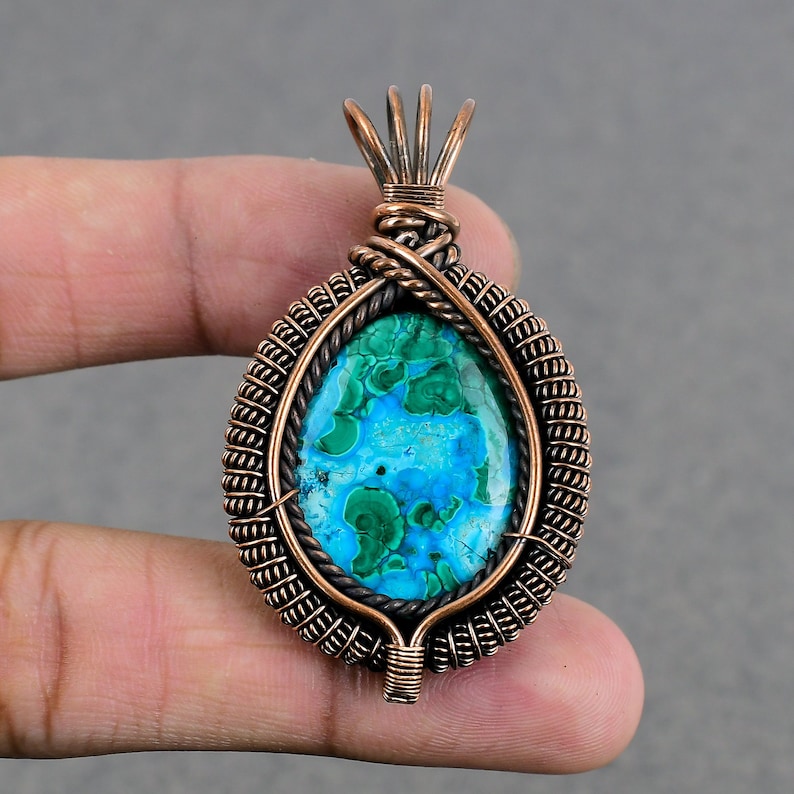 May include: A copper wire wrapped pendant featuring a green and blue malachite gemstone. The pendant has a woven wire design and a loop at the top for attaching a chain.