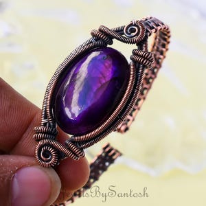 Puede incluir: Un brazalete envuelto en alambre de cobre con una gran piedra de ágata púrpura. El brazalete está hecho con un diseño de eslabón de cadena.