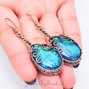 以下が含まれることがあります： 虹色の青と緑のダイクロイックガラスのカボションが付いた、銅線で巻かれたイヤリングのペア。イヤリングは繊細で複雑なデザインです。