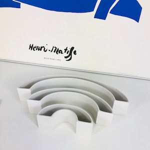 May include: A set of three white ceramic nesting bowls shaped like half-circles. The bowls are arranged in a stack, with the largest bowl on the bottom and the smallest bowl on top. The bowls are designed to be used as a decorative element or for serving snacks.