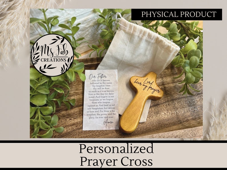 May include: A wooden prayer cross with the words "Lord hear my prayer" engraved on it. The cross is sitting on a wooden surface with a small white bag next to it. The cross is a light brown color and the words are in black.