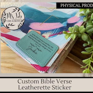 May include: A teal blue leatherette sticker with a bible verse and the name "Jackie" printed in black. The sticker is placed on a colorful abstract patterned journal.