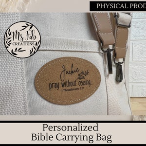 Op de afbeelding: Een witte canvas tas voor het dragen van een Bijbel met een bruine leren riem en een kurkachtige patch met de tekst "Jackil pray without ceasing... 1 Thessalonicenzen 5:17".