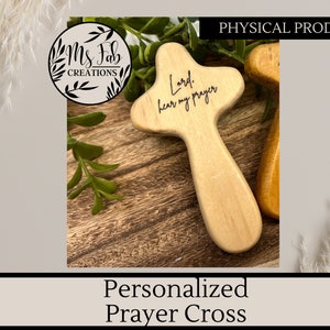May include: A small wooden cross with the words "Lord, hear my prayer" engraved on it. The cross is a light brown color and has a smooth finish. It is a personalized prayer cross.