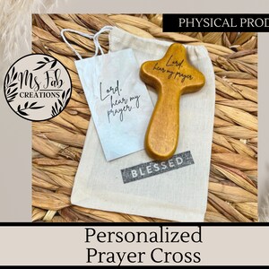 May include: A small wooden prayer cross with the words "Lord, hear my prayer" engraved on it. The cross is sitting on a white cloth bag with the word "BLESSED" printed on it.