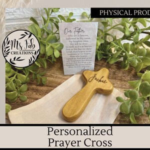 May include: A personalized wooden prayer cross with the name "Jackie" engraved on it. The cross is sitting on a white cloth bag with a small card that reads "Our Father, who art in heaven..." The cross is a light brown color and has a simple design.