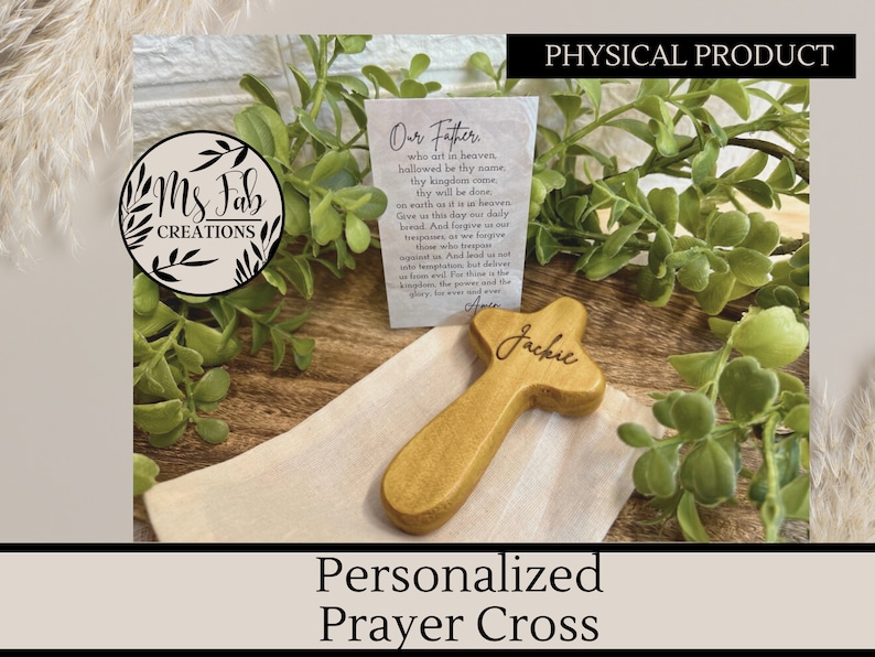 May include: A personalized wooden prayer cross with the name "Jackie" engraved on it. The cross is sitting on a white cloth bag with a small card that reads "Our Father, who art in heaven..." The cross is a light brown color and has a simple design.