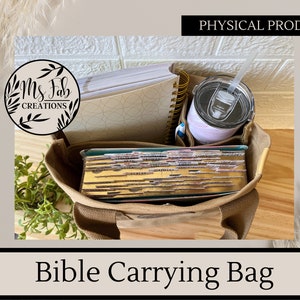 May include: A tan canvas tote bag with a zippered top, designed to carry a Bible, notebooks, and a tumbler. The bag is filled with a stack of books, a spiral notebook, and a tumbler with a straw. The bag has the text "Ms. Fab Creations" on a circular label.