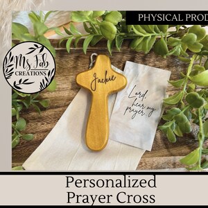 May include: A wooden prayer cross with the name "Jackie" engraved on it. The cross is light brown and has a small white cloth bag with the text "Lord, hear my prayer..." printed on it.