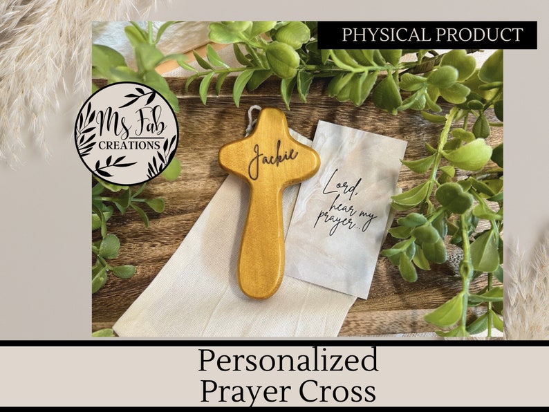 May include: A wooden prayer cross with the name "Jackie" engraved on it. The cross is light brown and has a small white cloth bag with the text "Lord, hear my prayer..." printed on it.