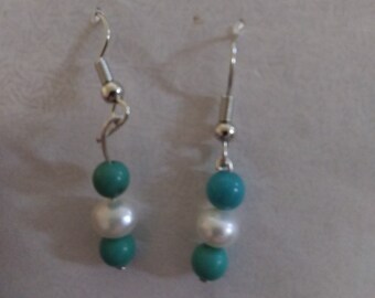 Op de afbeelding: Een paar oorhangers met elk drie kralen. De bovenste en onderste kralen zijn turquoise blauw en de middelste kraal is wit.