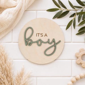 Może przedstawiać: Okrągły drewniany znak z napisem "IT'S A BOY" w zielonym kolorze. Znak otoczony jest elementami dekoracyjnymi, w tym gałązką oliwną, trawą pampasową i drewnianą zabawką. Ogłoszenie o narodzinach dziecka.