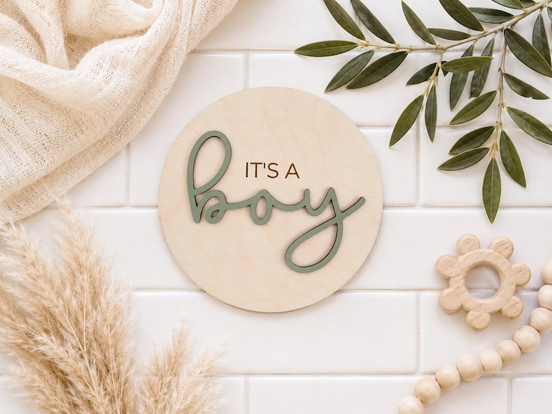 Może przedstawiać: Okrągły drewniany znak z napisem "IT'S A BOY" w zielonym kolorze. Znak otoczony jest elementami dekoracyjnymi, w tym gałązką oliwną, trawą pampasową i drewnianą zabawką. Ogłoszenie o narodzinach dziecka.