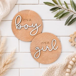 Może przedstawiać: Dwie okrągłe drewniane tabliczki z napisami &bdquo;It's a boy&rdquo; i &bdquo;It's a girl&rdquo; w białym, ozdobnym kroju pisma. Tabliczki są jasnobrązowe i leżą na białej, kafelkowej powierzchni, z elementami dekoracyjnymi.