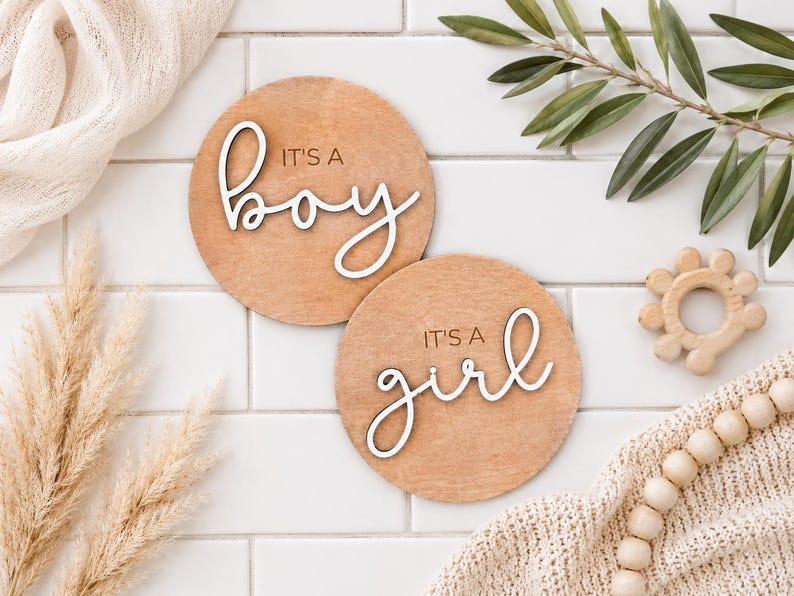 Może przedstawiać: Dwie okrągłe drewniane tabliczki z napisami &bdquo;It's a boy&rdquo; i &bdquo;It's a girl&rdquo; w białym, ozdobnym kroju pisma. Tabliczki są jasnobrązowe i leżą na białej, kafelkowej powierzchni, z elementami dekoracyjnymi.