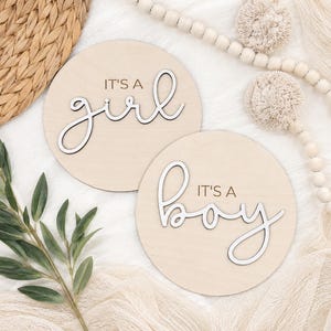 Może przedstawiać: Dwa okrągłe drewniane znaki z napisami "It's a girl" i "It's a boy" w kursywie. Znaki są jasnobeżowe z białymi literami. Tło zawiera elementy dekoracyjne.