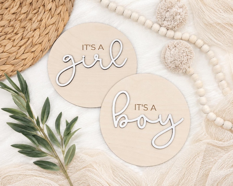 Może przedstawiać: Dwa okrągłe drewniane znaki z napisami "It's a girl" i "It's a boy" w kursywie. Znaki są jasnobeżowe z białymi literami. Tło zawiera elementy dekoracyjne.
