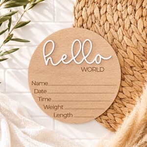 Może przedstawiać: Okrągły drewniany znak ogłoszenia o narodzinach dziecka z napisem "hello WORLD" w białym skrypcie. Znak ma linie na Imię, Datę, Godzinę, Wagę i Długość. Znak jest jasnobrązowy i otoczony elementami dekoracyjnymi.