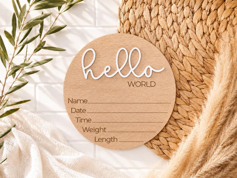 Może przedstawiać: Okrągły drewniany znak ogłoszenia o narodzinach dziecka z napisem "hello WORLD" w białym skrypcie. Znak ma linie na Imię, Datę, Godzinę, Wagę i Długość. Znak jest jasnobrązowy i otoczony elementami dekoracyjnymi.