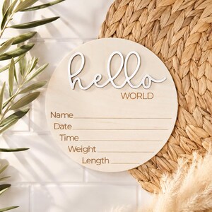 Może przedstawiać: Okrągły drewniany znak z napisem "hello WORLD" w białym skrypcie. Znak ma miejsce na wpisanie imienia, daty, godziny, wagi i wzrostu dziecka. Znak jest z jasnego drewna i jest umieszczony na tle z plecionki.