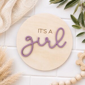 Może przedstawiać: Okrągły drewniany znak z napisem "IT'S A girl" w fioletowym skrypcie. Znak otoczony jest neutralnymi dekoracjami, w tym beżowym kocem, suszoną trawą pampasową i drewnianą grzechotką.