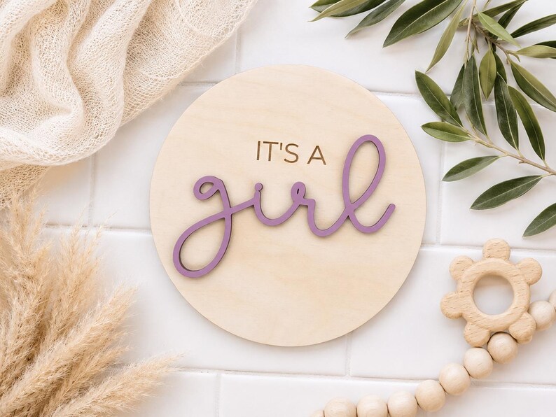 Może przedstawiać: Okrągły drewniany znak z napisem "IT'S A girl" w fioletowym skrypcie. Znak otoczony jest neutralnymi dekoracjami, w tym beżowym kocem, suszoną trawą pampasową i drewnianą grzechotką.