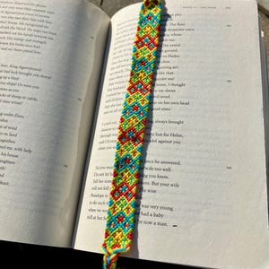 May include: A colorful friendship bracelet with red, yellow, blue, and green threads woven together in a geometric pattern. The bracelet is lying on top of an open book.