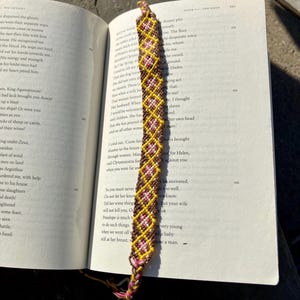 May include: A brown, yellow, and pink friendship bracelet with a diamond pattern. The bracelet is lying on an open book.