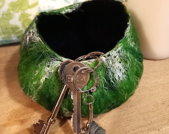 May include: A handmade, green felt bowl with a black interior, holding a set of keys. The bowl has a textured, organic appearance with white accents. A silver butterfly charm hangs from the key ring. The bowl is a decorative item for storing small objects.