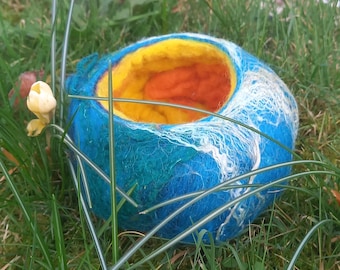 May include: A handmade felted bowl in shades of blue, teal, yellow, and orange, resting in green grass. The bowl has a rounded shape with a textured surface and a contrasting interior. White fibers are incorporated into the blue exterior.